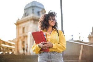 a woman in a yellow jacket and light grey distressed jeans, holding a red register in front of a landmark building, epitomizing the essence of a modern-day international student and F-1 visa holder.